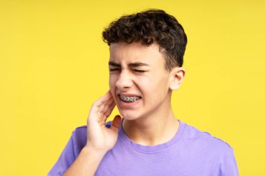 Yüzüne dokunan diş teli takan üzgün genç bir çocuk. Gözleri kapalı diş ağrısı çekiyor. Sarı arka planda izole edilmiş bir genç. Tedavi kavramı, diş sağlığı