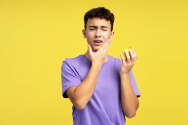 Dişleri birbirine değen mutsuz mutsuz bir ergen, sarı arka planda kapalı duran bir elmayla diş ağrısı çekiyor. Diş ve tedavi kavramı