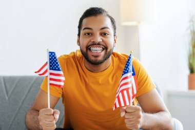 Gülümseyen bir Afro-Amerikan futbol fanatiğinin portre fotoğrafı, diş telleriyle, bir kanepede oturuyor, ABD bayraklarını tutuyor ve kameraya bakıyor.