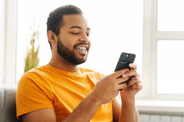 Fotoğrafta diş teli takmış gülümseyen, Afro-Amerikan bir adam var. Kanepede rahatça oturuyor ve cep telefonuyla sohbet ediyor.