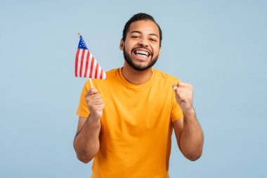 Diş teli takan, Amerikan bayrağı tutan, yumruklarını sıkarak 'evet' diyen ve mavi arka planda izole edilmiş kameraya bakan mutlu Afrikalı Amerikalı bir adam. Seçim sonuçları konsepti bekleniyor