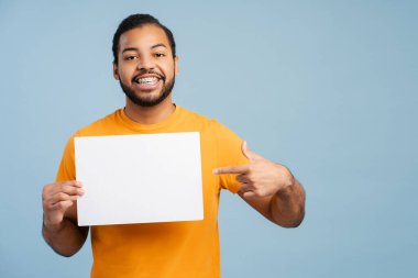 Diş telleri boş bir tabela tutan mutlu Afrikalı Amerikalı adam mavi arka planda izole edilmiş kameraya bakarken parmağıyla işaret ediyor. Model konsepti