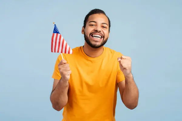 Happy African American man with dental braces, holding American flag, making a \'yes\' gesture with clenched fists, and looking at camera, isolated on blue background. Waiting election results concept