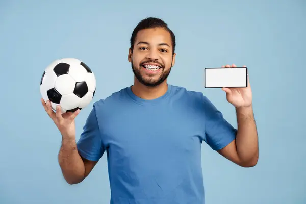 Radosny Afroamerykanin Aparatem Zębach Trzymający Piłkę Nożną Telefon Komórkowy Pokazujący — Zdjęcie stockowe