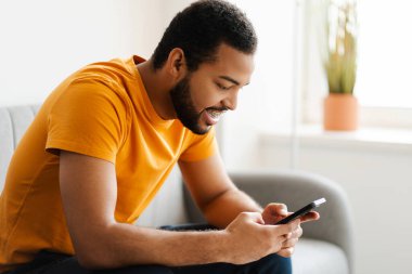 Dişlerinde diş teli olan gülümseyen Afro-Amerikan bir adam cep telefonu tutuyor, internetten iletişim kuruyor rahat koltukta mesaj okuyor, evde dinleniyor. Teknoloji, sosyal medya kavramı 