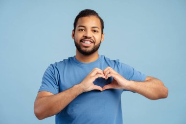 Mavi arka planda izole edilmiş aboneler için kalp şekli gösteren diş telleri olan mutlu Afrikalı Amerikalı adam. Pozitif yaşam tarzı, sosyal medya kavramı 