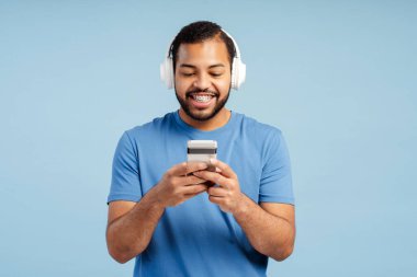 Kulaklık takan, cep telefonu tutan, mavi arka planda izole edilmiş müzik dinleyen gülümseyen yakışıklı adamın portresi. Eğlence konsepti, teknoloji. 
