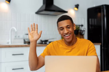 Çekici gülümseyen Afrikalı Amerikalı adam el sallıyor, video konferansı yapıyor, dizüstü bilgisayar kullanıyor, evde çalışan serbest çalışan, mutfakta oturan. Kendine güvenen öğrenci çalışıyor. Çevrimiçi eğitim kavramı