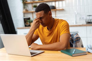 Depresyondaki üzgün Latin adam evde çalışıyor, dizüstü bilgisayar kullanıyor, gözleri kapalı, migreni var, baş ağrısı var, kötü haberler var. Mutsuz hipster yöneticisi masasında oturuyor. Uzak iş kavramı, sağlık hizmetleri