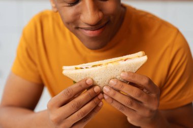 Yakışıklı Afro-Amerikan bir adamın elinde sandviçle evdeki mutfakta otururken yemek yiyip eğlenen portresi, yakın plan. Öğle yemeği, yemek, akşam yemeği kavramı
