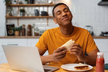Yakışıklı, mutlu Latin adam lezzetli sandviçler tutuyor, yemek yiyor, kapalı gözlerle evde oturmuş eğleniyor. Yemek kavramı, evde çalışan serbest çalışan biri.
