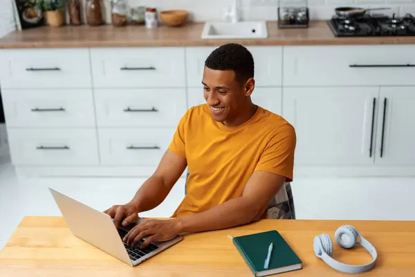 Portrait of smiling African American man, freelancer, copywriter typing on keyboard wearing stylish yellow t shirt sitting on workplace, using laptop, remote job. Online education concept