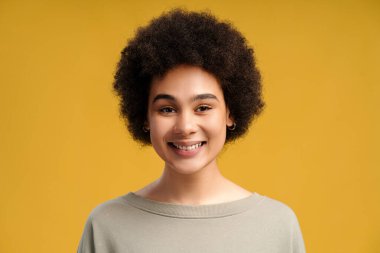 Kıvırcık saçlı, gülümseyen, güzel Afro-Amerikan kadın sarı arka planda tek başına duran kameraya bakıyor. Doğal güzellik kavramı, kozmetoloji