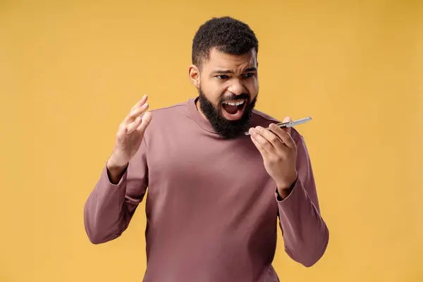 stock image Angry, pensive, bearded African American man using mobile phone, recording voice message, shouting standing isolated on yellow background. Communication concept