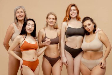 Çok ırklı, gülümseyen, şık sutyen ve külot giyen ve kameraya bakan bir grup güzel kadın. Bej arka planda izole edilmiş kadınlar stüdyoda birlikte poz veriyorlar. Uluslararası Kadınlar Günü konsepti