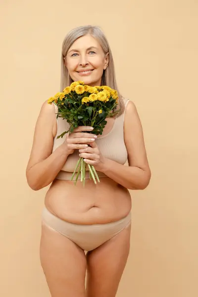 Beautiful senior 60 year old woman wearing stylish lingerie holding beautiful bouquet of chrysanthemums looking at camera standing isolated on beige background. Concept of celebration, birthday