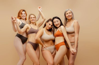 Bir grup çok ırklı, gülen kadın, şık seksi iç çamaşırları giyen, kameraya bakan, bej arka planda, stüdyoda poz veren çekici mankenler. Reklam konsepti, kadınlık