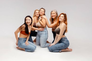 Bir grup gülümseyen, çok ırklı kadın şık sutyenler ve kot pantolonlar giyip kameraya bakarak oturuyorlar. Stüdyoda beyaz arka planda poz veren mankenler. Reklam konsepti, doğal güzellik.