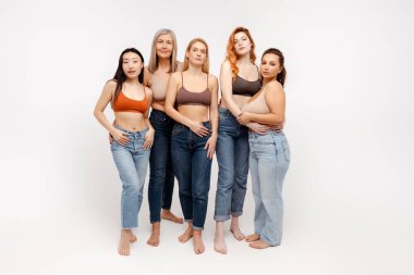 Bir grup çekici, güler yüzlü, çok kültürlü kadın şık kıyafetler, sutyen ve kot pantolon giyerek beyaz arka planda izole bir şekilde kameraya bakıyorlar. Doğal güzellik kavramı