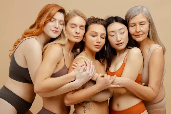 Group of attractive cheerful multiracial women wearing stylish lingerie embracing with closed eyes. Beautiful fashion models posing isolated on beige background. Concept natural beauty, diversity