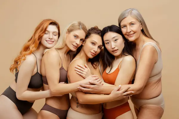 Group of smiling multiracial women wearing stylish lingerie embracing looking at camera. Beautiful fashion models posing isolated on beige background. Concept natural beauty, diversity