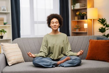 Çekici, güzel, sıradan kıyafetler giyen Afro-Amerikan kadın Lotus pozisyonunda rahat bir koltukta oturuyor ve evde meditasyon yapıyor. Uyum kavramı, denge