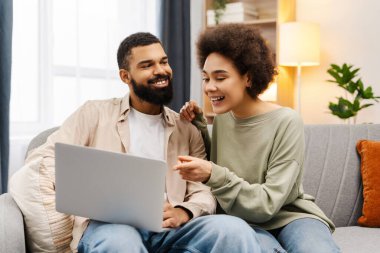 Çekici gülümseyen Afro-Amerikan çiftin portresi, dizüstü bilgisayar kullanan, evde çalışan, rahat koltukta otururken online alışveriş yapan yakışıklı erkek ve kadın. Teknoloji konsepti, uzak meslek