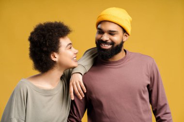 Çekici Afro-Amerikan sakallı erkek ve kadın kıvırcık saçlı, şık kıyafetler giyen, sarı şapkalı, sarılan, konuşan, sarı arka planda tek başına duran. Aşk kavramı, iletişim.