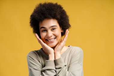 Sarı arka planda izole edilmiş kameraya bakan kıvırcık saçlı, gülümseyen Afrikalı Amerikalı kadının portresi. Cilt bakımı kavramı, doğal güzellik.