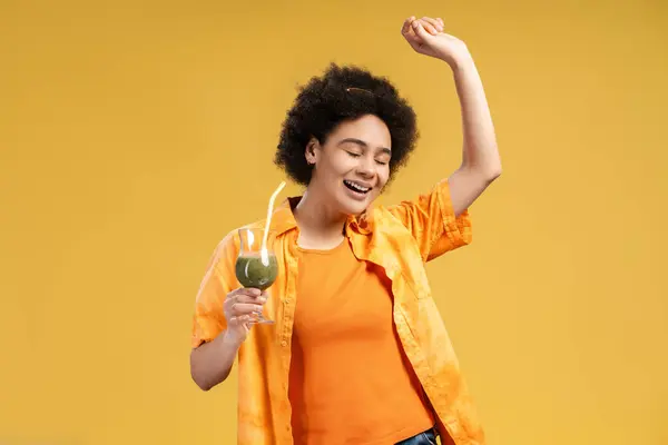 Excited, smiling African American woman holding cocktail with eyes closed dancing, having fun isolated on yellow background. Beautiful female wearing orange shirt. Concept of summer, party celebration