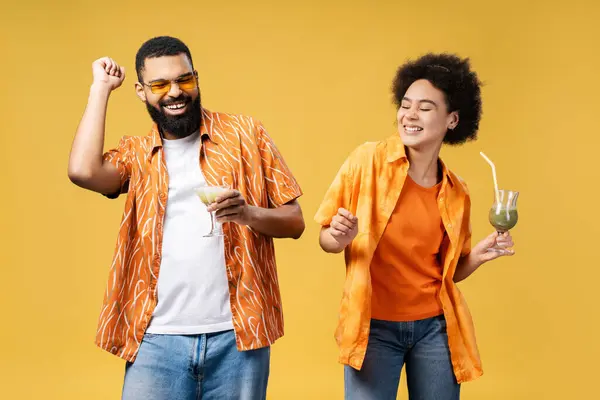 stock image Attractive, happy African American couple holding cocktails having fun isolated on yellow background. Smiling man in sunglasses and woman dancing relaxing with closed eyes. Party, celebration concept