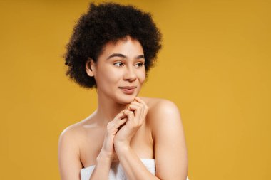 Beyaz havlu giymiş kıvırcık saçlı neşeli Afro-Amerikan kadın başka tarafa bakıyor. Sarı arka planda izole edilmiş güzel bir kadın model. Cilt bakımı kavramı, kozmetik prosedürleri