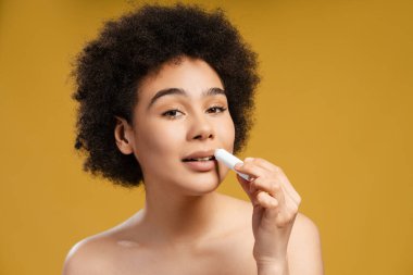 Güzel, kıvırcık saçlı, ruj süren, aynaya bakan, sarı arka planda tek başına duran Afro-Amerikan bir kadın. Koruma kavramı, kozmetik ürün