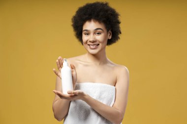Beyaz havluyla duştan sonra beyaz havluyla kıvırcık saçlı, gülümseyen Afro-Amerikan genç kadın elinde beyaz şişeyle kameraya bakıyor. Sabah rutini kavramı, kozmetik ürünü, reklam