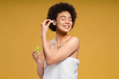 Duş aldıktan sonra beyaz havluyla mutlu Afro-Amerikalı kadın duş üstüne serum uyguluyor. Sabah rutini, gözleri kapalı sarı arka planda tek başına duruyor. Kozmetoloji kavramı
