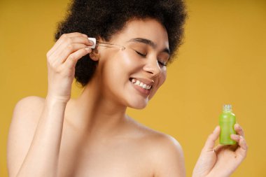 Kıvırcık saçlı, yeşil şişeli, yüzüne yağ süren, gözleri kapalı beyaz bir havlu giyen, sarı arka planda izole bir şekilde duran, kıvırcık saçlı, genç Afrikalı bir kadın. Kozmetoloji kavramı