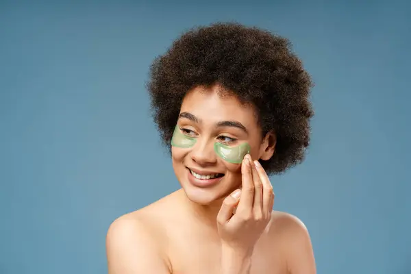 Authentic, happy African American model, woman with curly hair with eye patches looking away on copy space, standing isolated on blue background. Concept of skin care, cosmetic product