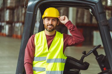 Gülümseyen, sakallı adam, şoför, kask takan, forklift yanında duran, kameraya bakan, depoda duran. Lojistik kavramı, dağıtım