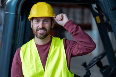 Yakışıklı, sakallı adam, şoför, kask takıyor ve forklift yanında duruyor, kameraya bakıyor, depoya yakın duruyor. Lojistik kavramı, dağıtım