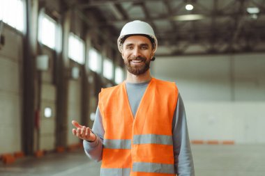 Başarılı, sakallı, profesyonel ustabaşı koruyucu beyaz kask takıyor ve bir şeyleri açıklıyor, el kol hareketi yapıyor, depoda duran kameraya bakıyor. Endüstri kavramı