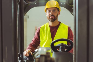 Gülümseyen, yakışıklı, sakallı bir adam, kasketli bir kameraman, sürücü arabası, direksiyonda forkliftler, kameraya bakıyor. Dağıtım ve ulaşım kavramı
