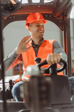 Yakışıklı, gülümseyen adam, forkliftte oturan ustabaşı, elini sallıyor, başka tarafa bakıyor, selamlıyor, sıkı şapka ve iş kıyafeti giyiyor. Depo işi, ulaşım, araç kavramı