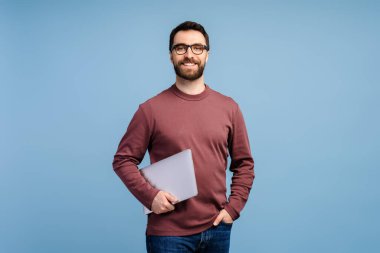 Çekici, gülümseyen, gözlüklü, elinde dizüstü bilgisayarla kameraya bakan, mavi arka planda tek başına duran sakallı bir adam. Eğitim kavramı, çevrimiçi teknoloji