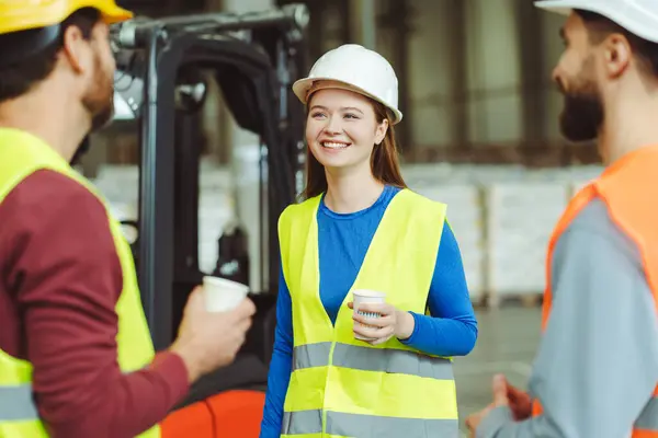 Beautiful, smiling, young woman in hard hat holding cup of coffee, looking away, listening to her colleagues, talking, standing in warehouse, coffee break. Concept of relaxation, logistics, industry