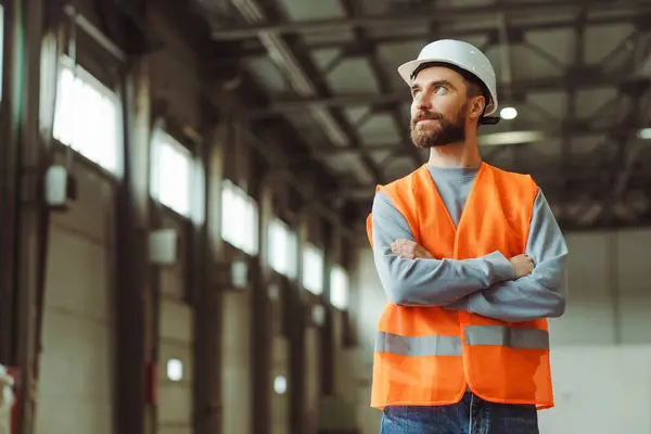 Attractive, handsome factory man wearing hard hat and workwear with crossed arms standing in warehouse, looking away at copy space. Concept of advertisement, engineer job