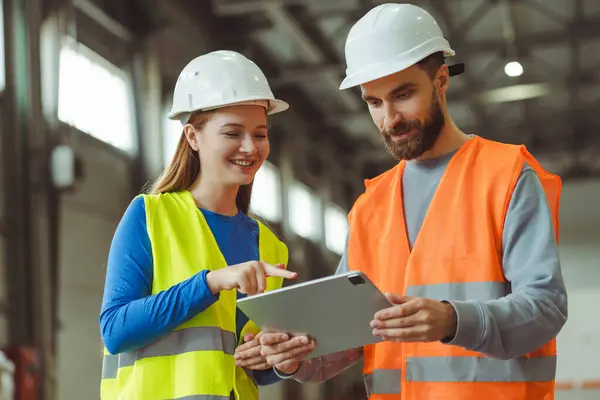 Happy engineers, factory foreman and female worker wearing protective white helmets, using digital tablet, planning startup, standing in warehouse. Cooperation, teamwork concept