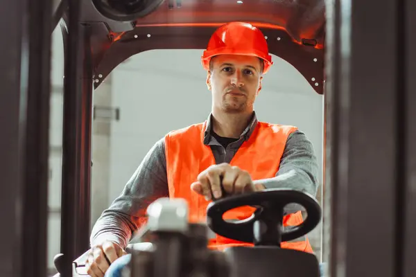 Serious, attractive factory man, driver wearing protective helmet and workwear, driving forklift, working in warehouse. Concept of logistics, industry, transportation