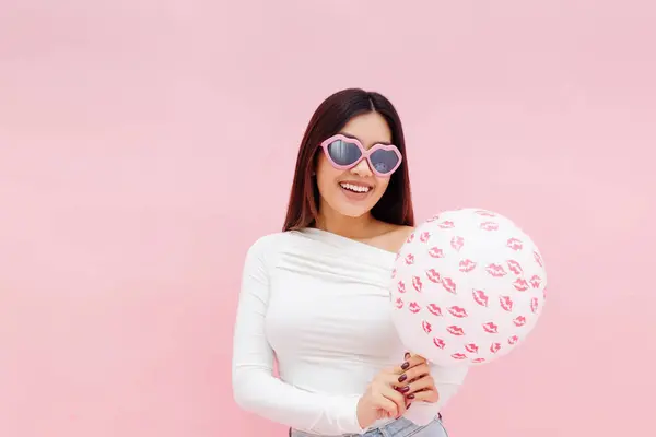 Happy smiling asian lady in eyeglasses holding balloon and looking at camera while posing isolated on pink background. Valentines day celebration concept