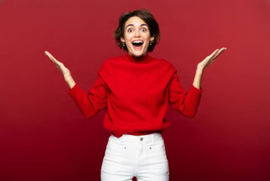 Şaşırmış bir kadının portresi şaşkınlıktan nefesi kesiliyor, ellerinin yanı başında duruyor, ağzını açık tutuyor, hayrete düşüyor. Kırmızı arkaplanda kapalı stüdyo çekimleri