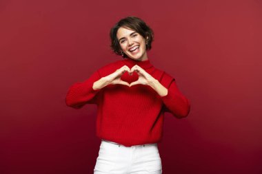 Portrait photo of joyful, attractive woman making a heart gesture with her hands, celebrating Valentine's Day, posing isolated on a red background, and looking into the camera  clipart